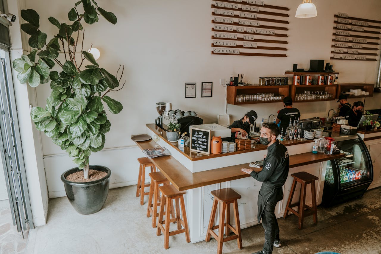 about-us-02 Chic café interior with a barista working next to a large potted plant, offering a welcoming ambiance.