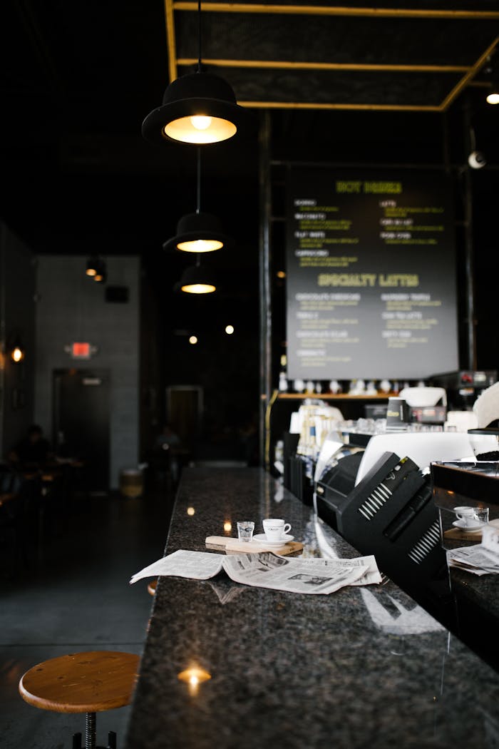 about-us-01 A cozy cafe in Greensboro featuring a granite bar with a cup, newspapers, and hanging lamps.